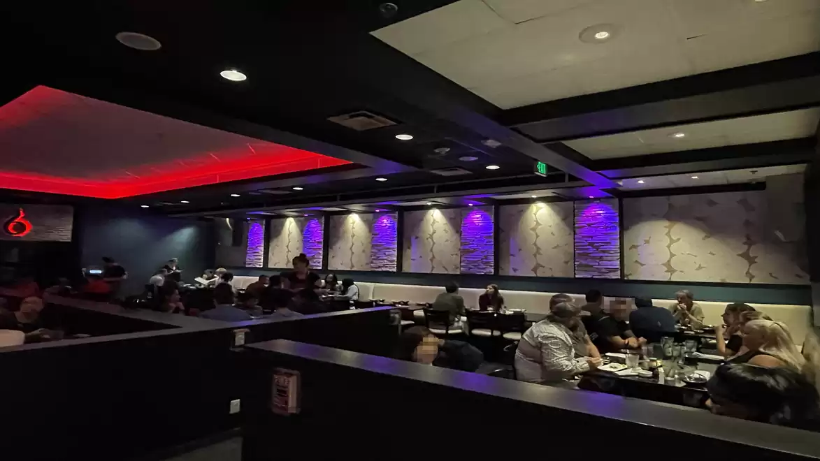 A modern restaurant with dark lighting and people dining at several tables. The walls are decorated with stylish patterns, and there is striking red and purple ceiling lighting.