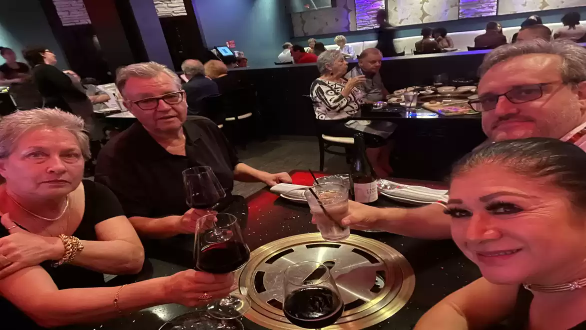 Four people sitting at a table in a restaurant, holding red wine glasses, with other guests visible in the background.