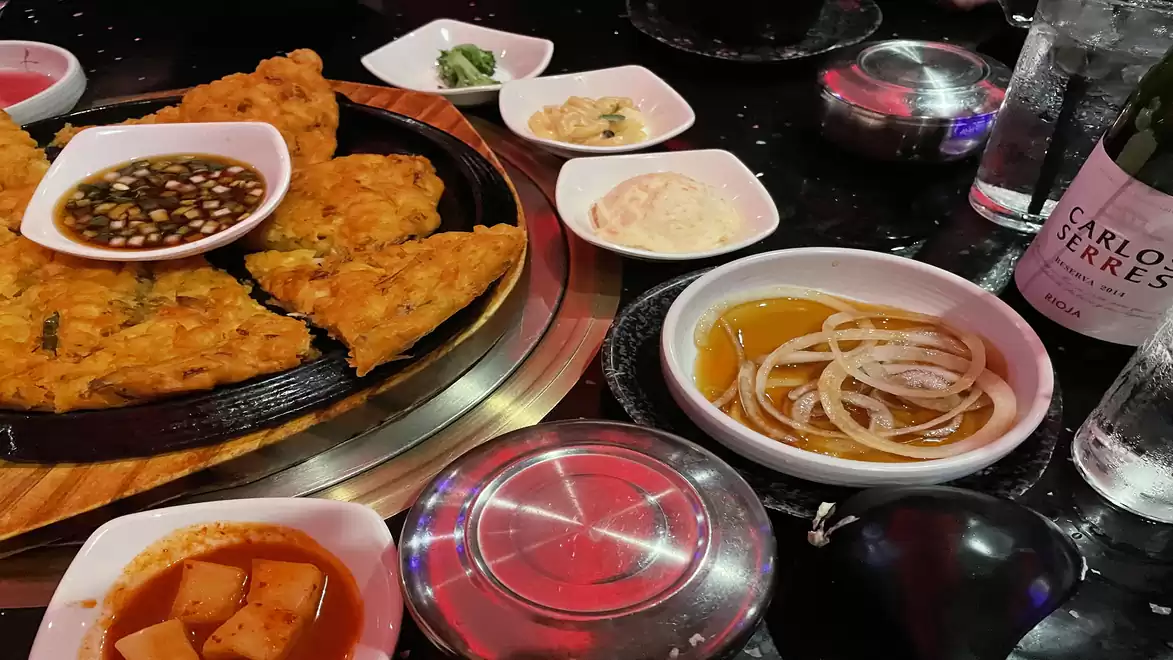 A table full of delicious dishes: crispy pancake with dip, bowls with various sauces, vegetables, and drinks.