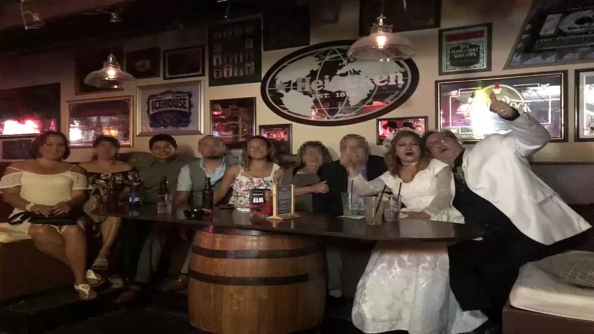 A group of people happily posing in a bar with atmospheric lighting. They are gathered around a table resembling a wooden barrel, with some holding drinks.