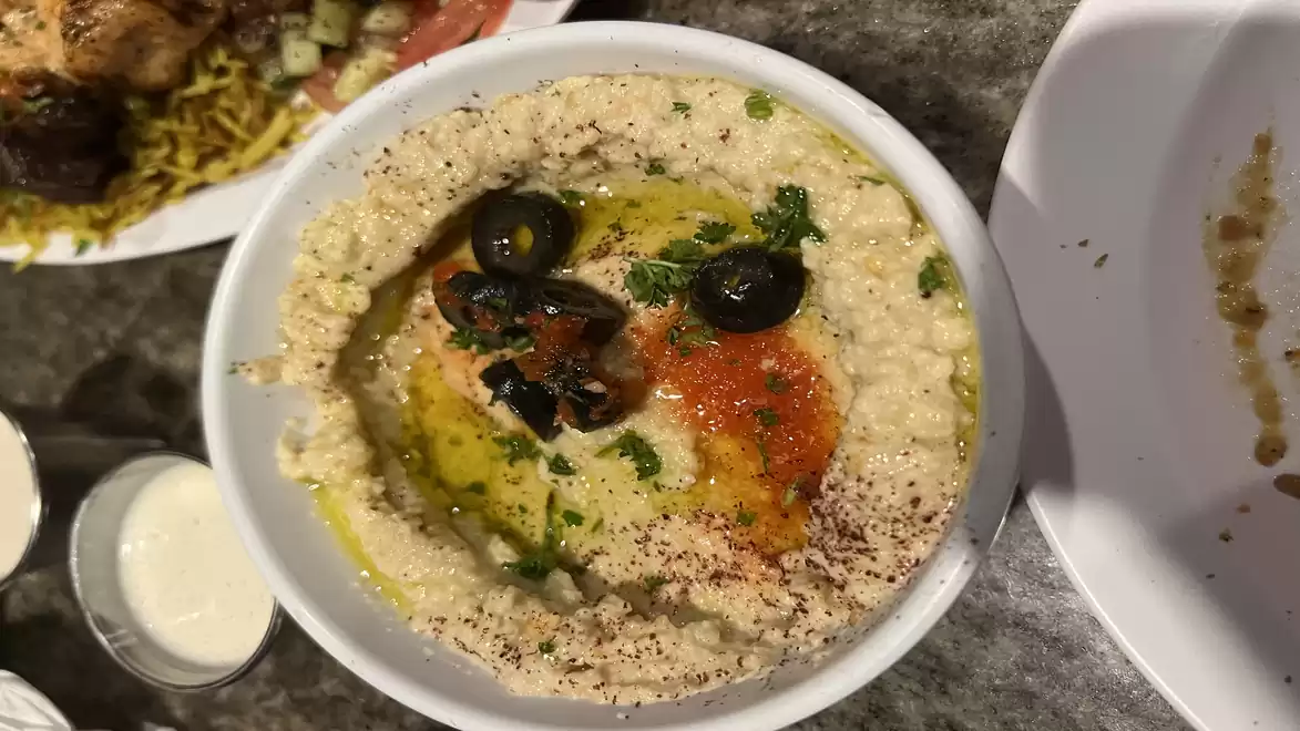 Close-up of a bowl of hummus with olives, spices, and oil, on a table with other dishes.