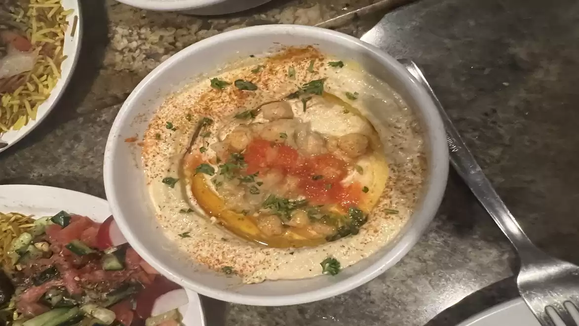 Close-up of a plate with hummus, garnished with olive oil, chickpeas, and spices, on a marble table with side dishes in the background.