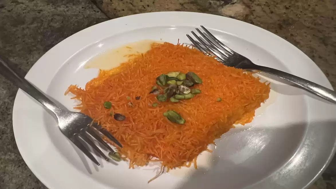 A plate with a piece of Kunafa, topped with orange threads and garnished with chopped pistachios, served on a white plate with two forks.