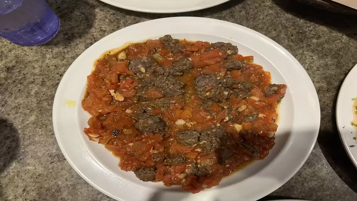 A plate with a dish of tomato sauce and meat pieces on a granite table, next to a glass with a beverage.