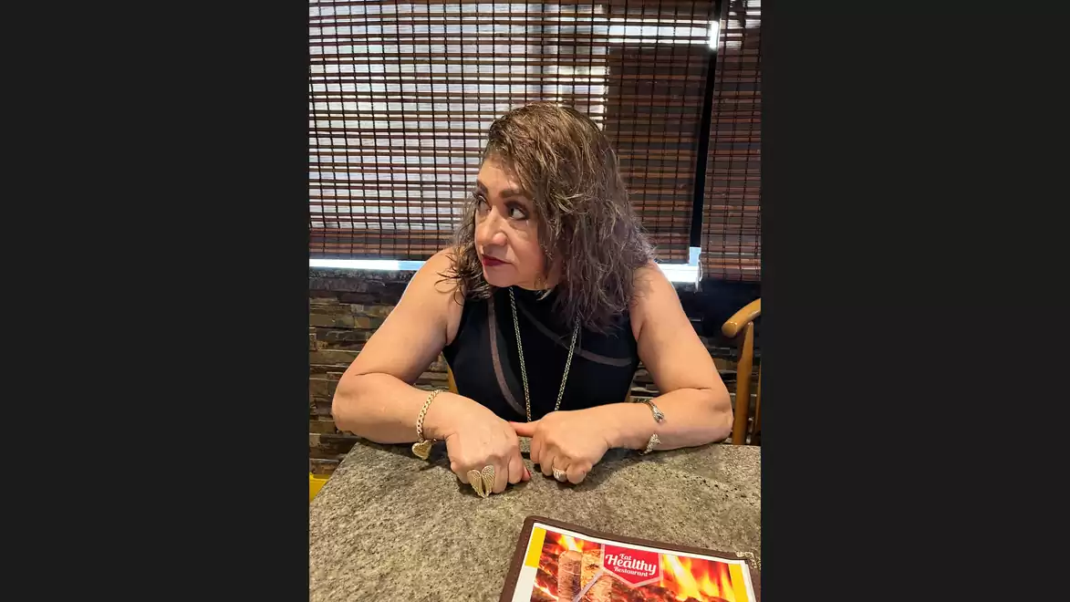 A woman with curly hair sits at a table in a restaurant and looks to the side.