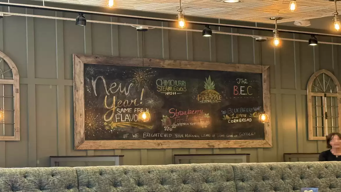 A cozy café interior with a decorative chalkboard on the wall featuring various handwritten menu options and specials. The chalkboard is surrounded by rustic wooden frames.