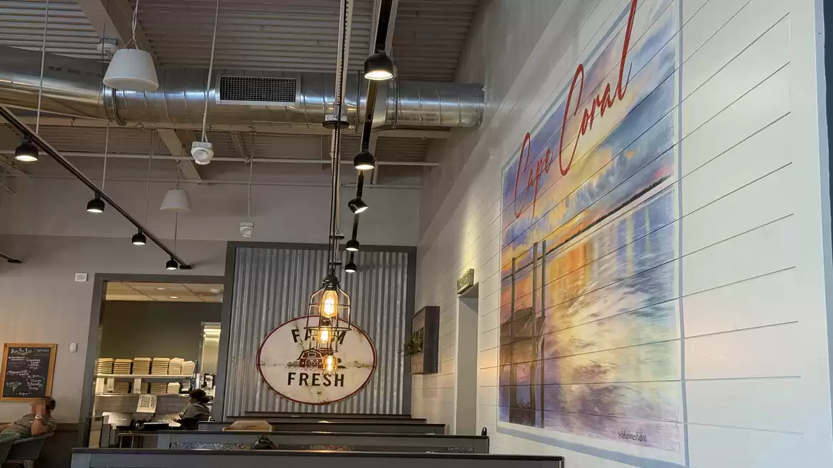Modern interior of a cafe with an industrial design, visible pipes on the ceiling, and stylish lamps. A large colorful painting decorates the white wall.