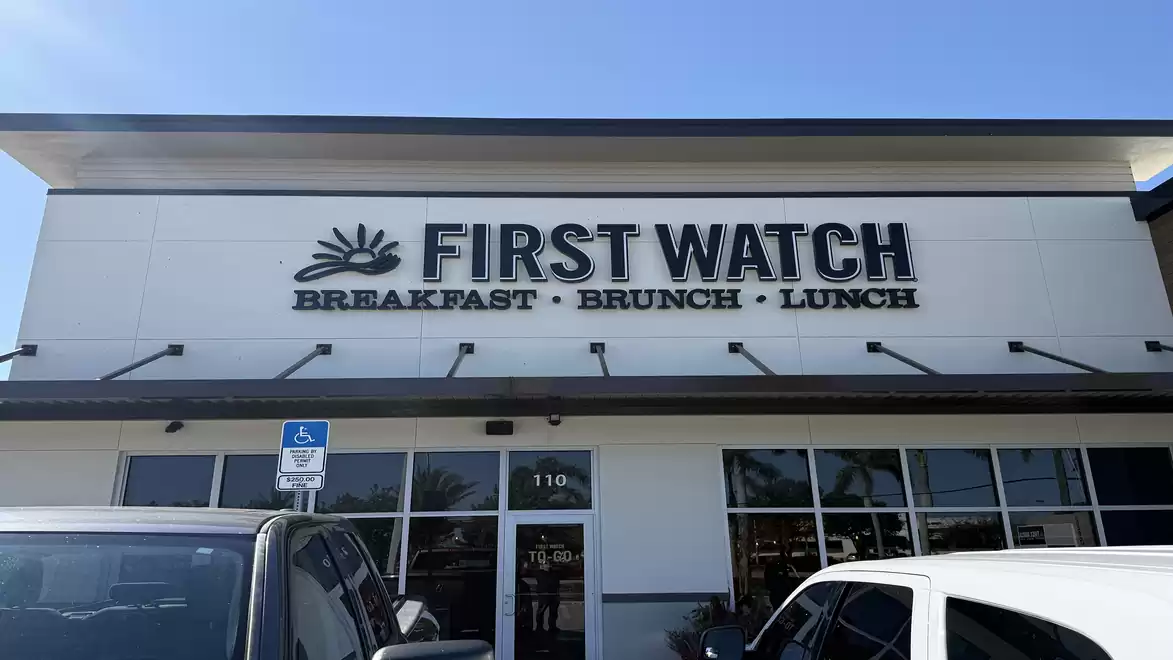 Exterior view of a restaurant named 'First Watch', specializing in breakfast, brunch, and lunch. The sky is clear and blue.