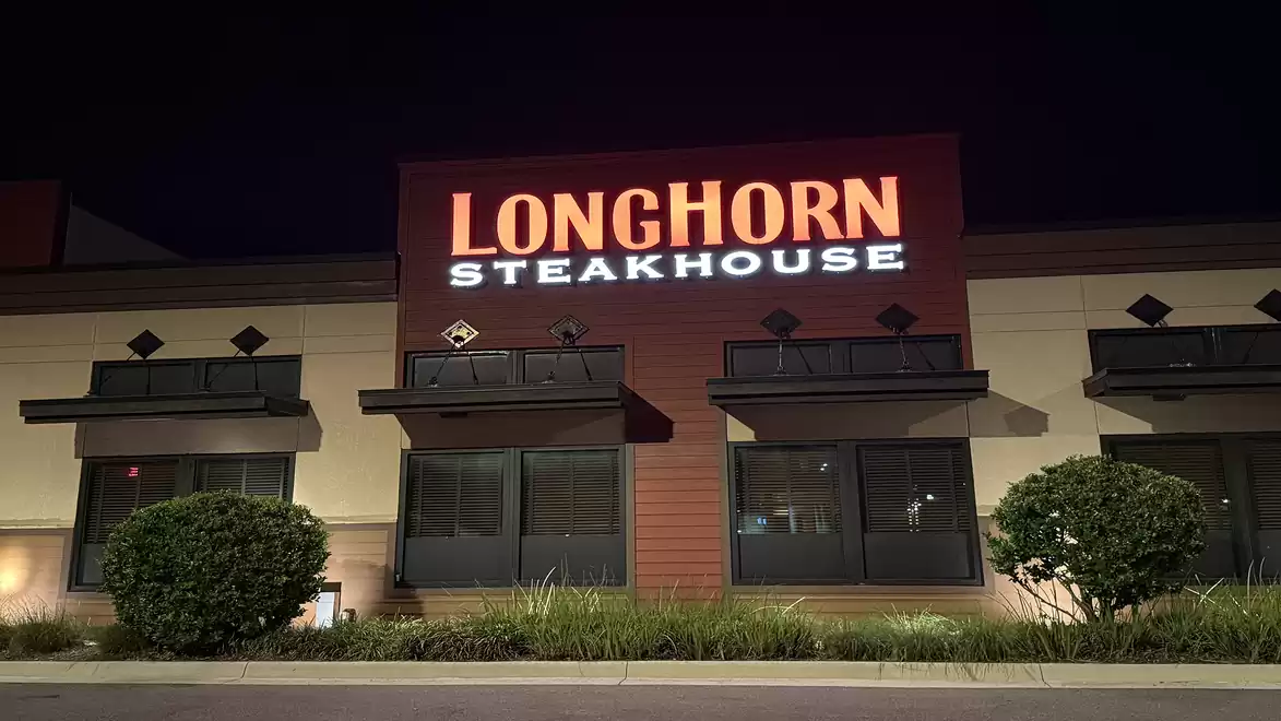 Exterior view of a LongHorn Steakhouse at night with illuminated logo and surrounding plants.