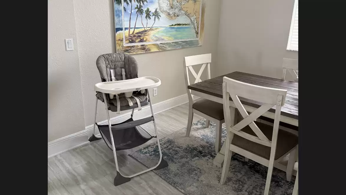 Dining room with a high chair and a dining table with four chairs. A tropical beach picture hangs on the wall.