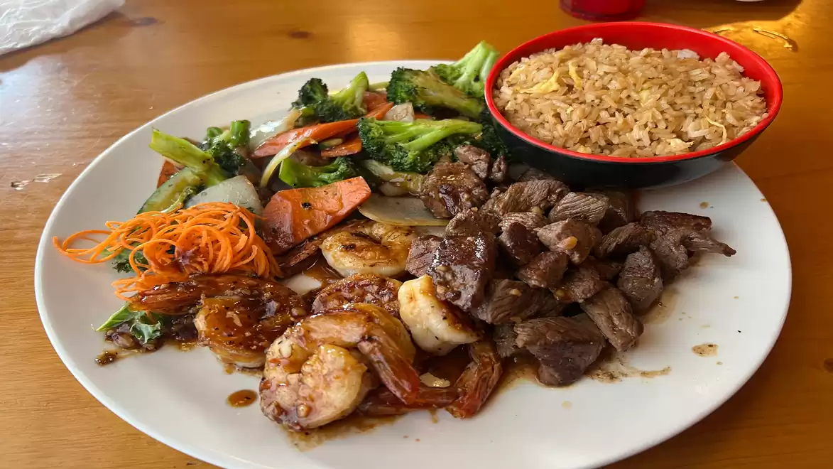 A plate with grilled shrimp, diced beef, stir-fried vegetables, and a bowl of rice on a wooden table.