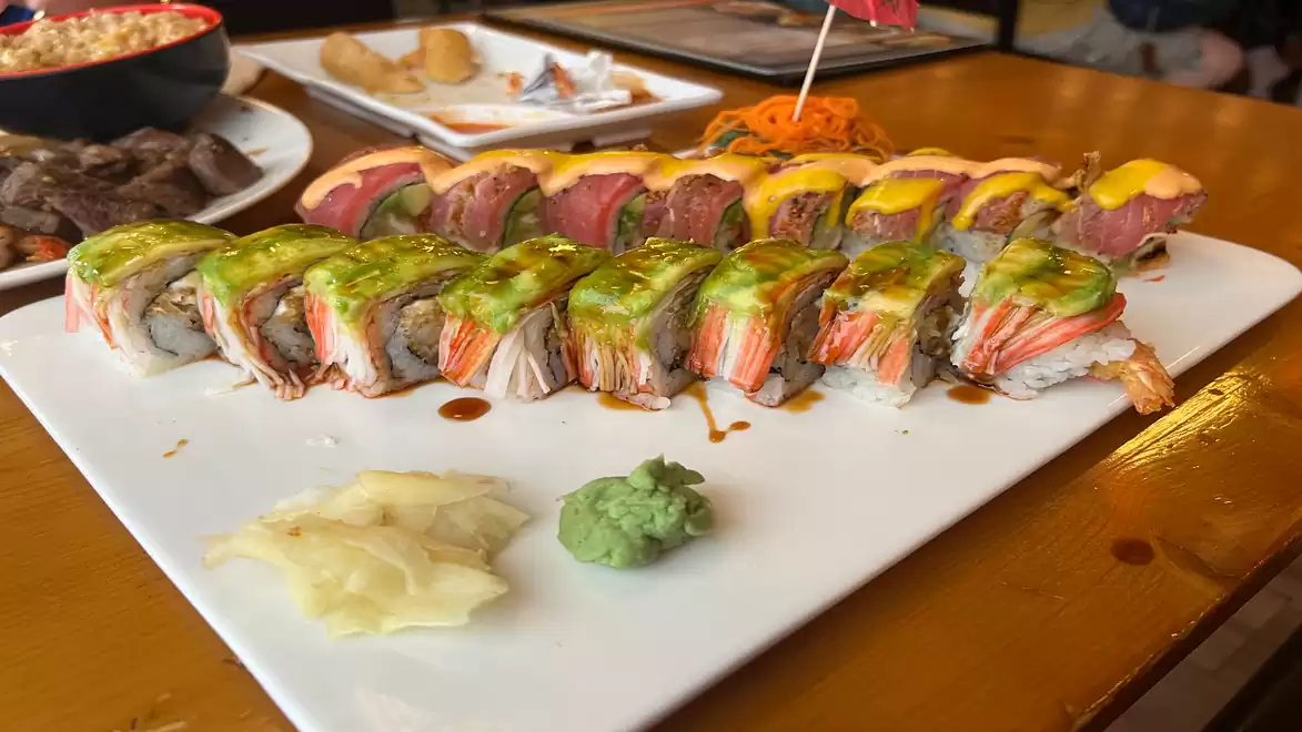 A variety of sushi rolls on a white plate, topped with avocado, raw fish, and a green dollop of wasabi. In the background, there are additional plates with different dishes.