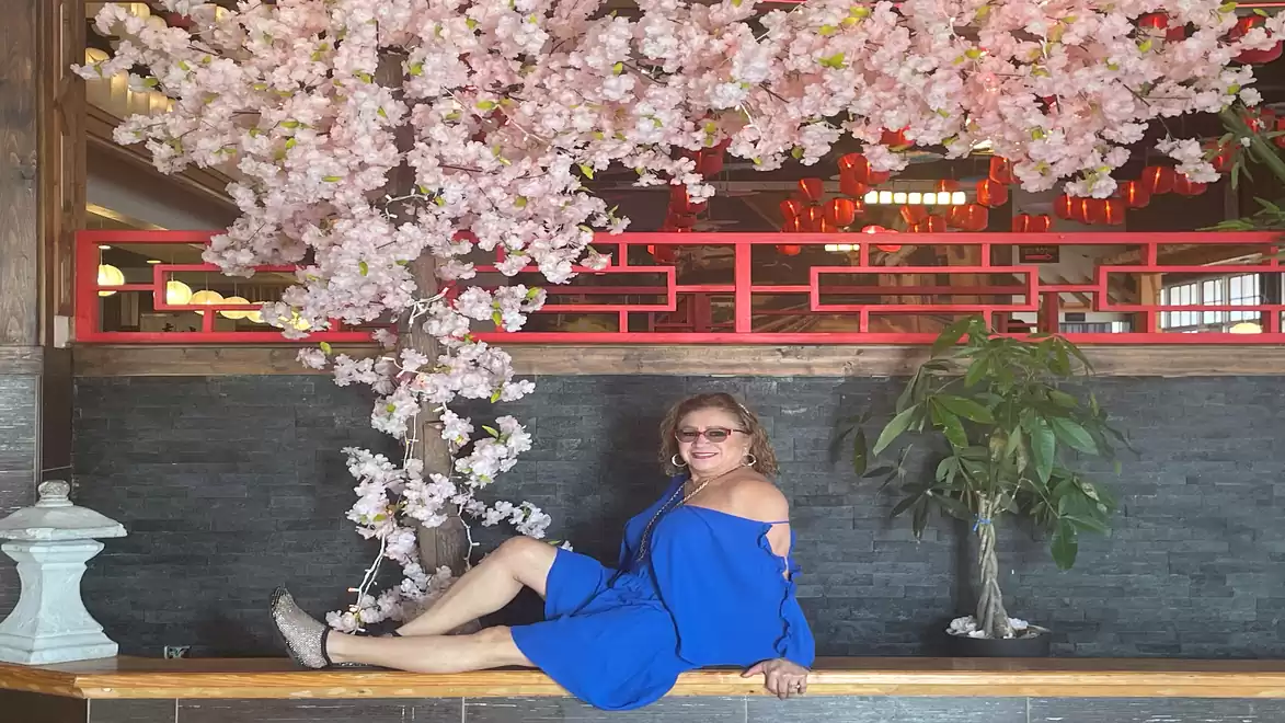 A woman in a blue dress sits under artistically arranged cherry blossoms in a stylish interior with oriental design elements.