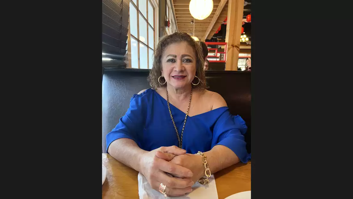 A woman in a blue top is sitting at a table in a restaurant, smiling at the camera. Windows and lighting are visible in the background.