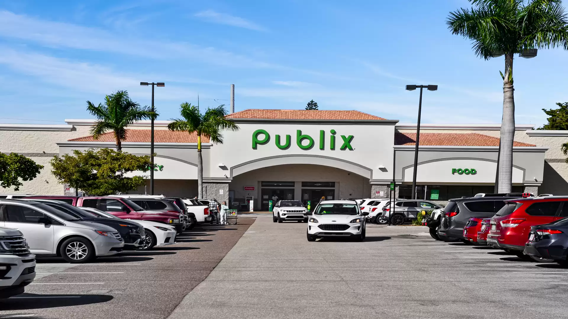 Entrance of a Publix supermarket with a parking lot, lined with palm trees and vehicles on a sunny day.