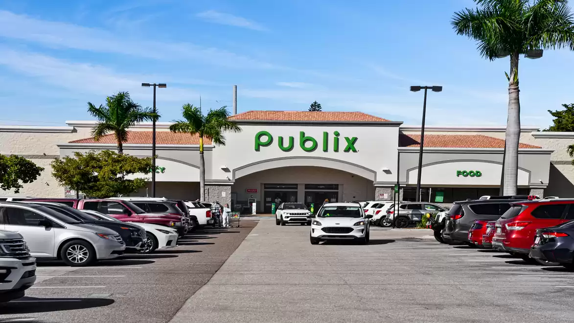 Entrance of a Publix supermarket with a parking lot, lined with palm trees and vehicles on a sunny day.