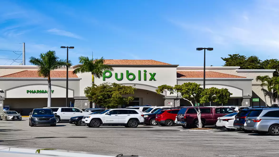 A parking lot with several cars in front of a building with the Publix sign. It's a sunny day with a clear blue sky, some palm trees are visible.