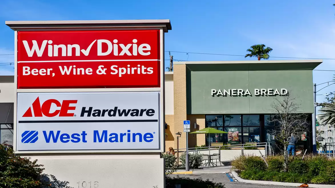 Store signs for Winn-Dixie, Ace Hardware, and West Marine in front of a Panera Bread in a sunny shopping area.