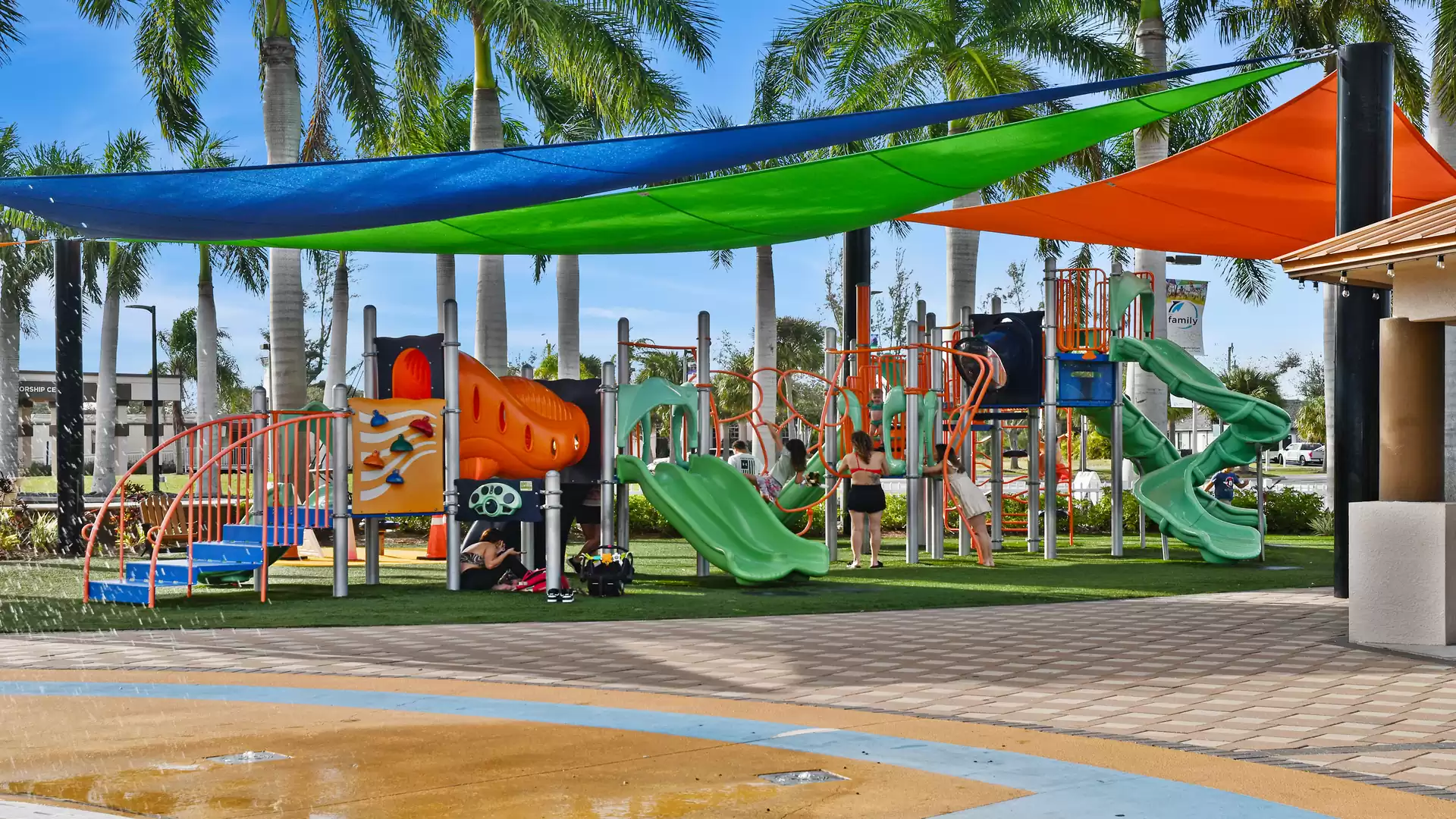 A colorful playground with climbing frames and slides, surrounded by palm trees and with canopies in different colors.
