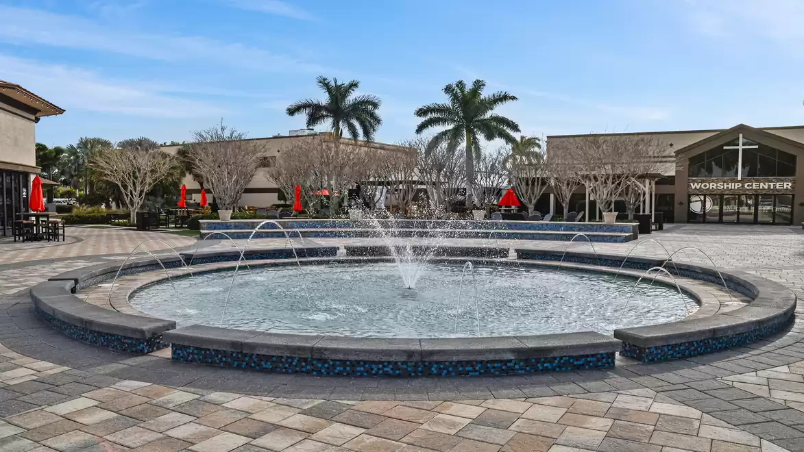 Fountain on a paved square with palm trees in the background and a modern building.