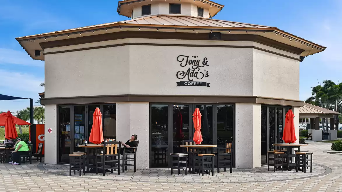 A modern café with an octagonal shape, large glass windows, and red umbrellas. It is located in a sunny setting.