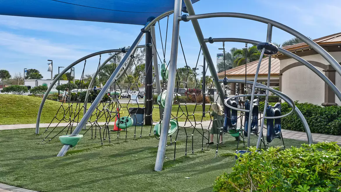 Modern playground with climbing structure and slides, sunny day in the park, surrounded by trees and buildings.