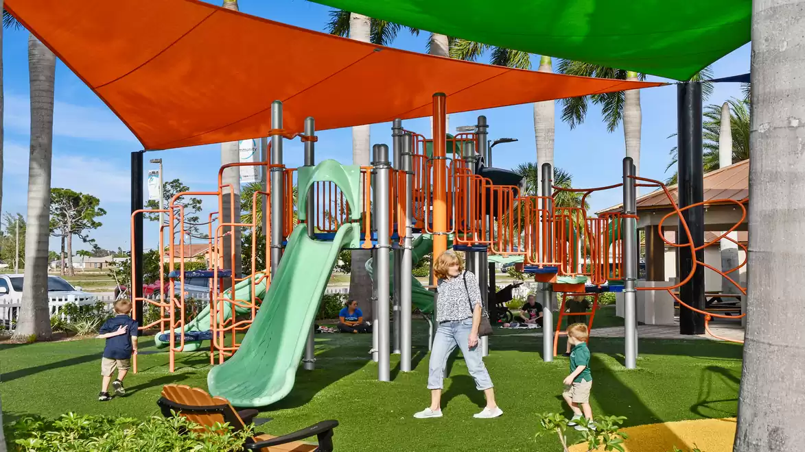 A modern playground with a green slide and orange sunshade. Children are playing on the climbing structure while an adult stands in the center. The area is surrounded by palm trees.