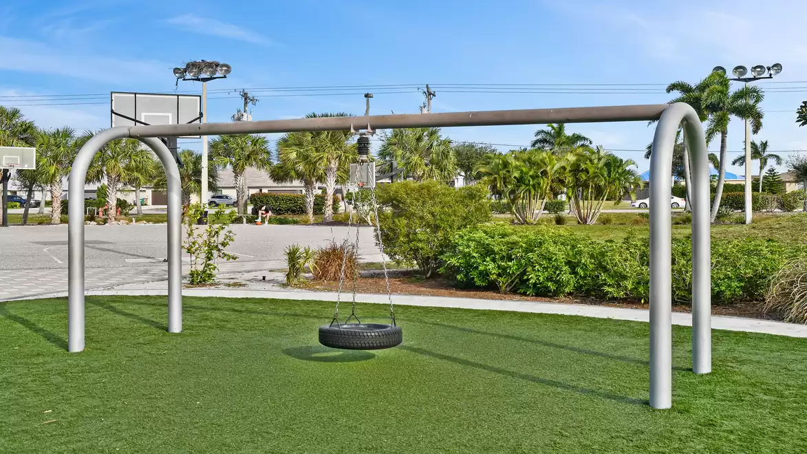 A playground with a large tire swing suspended from a metal frame, surrounded by green grass and trees.