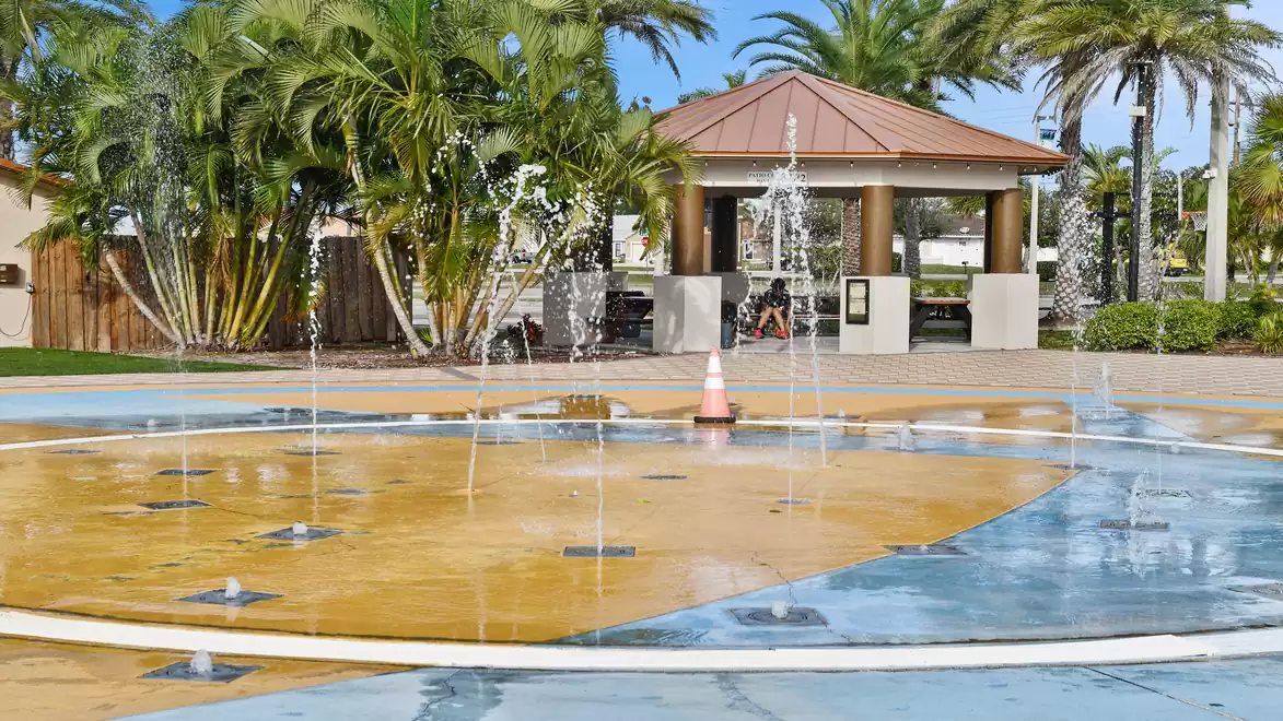 A round splash pad with fountains in front of a pavilion, surrounded by palm trees on a sunny day.
