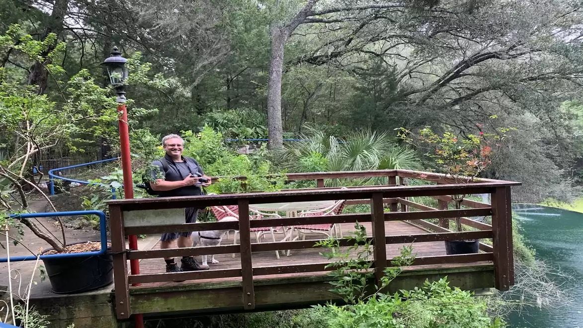 Man standing on a wooden deck surrounded by lush forest with garden furniture in the background.