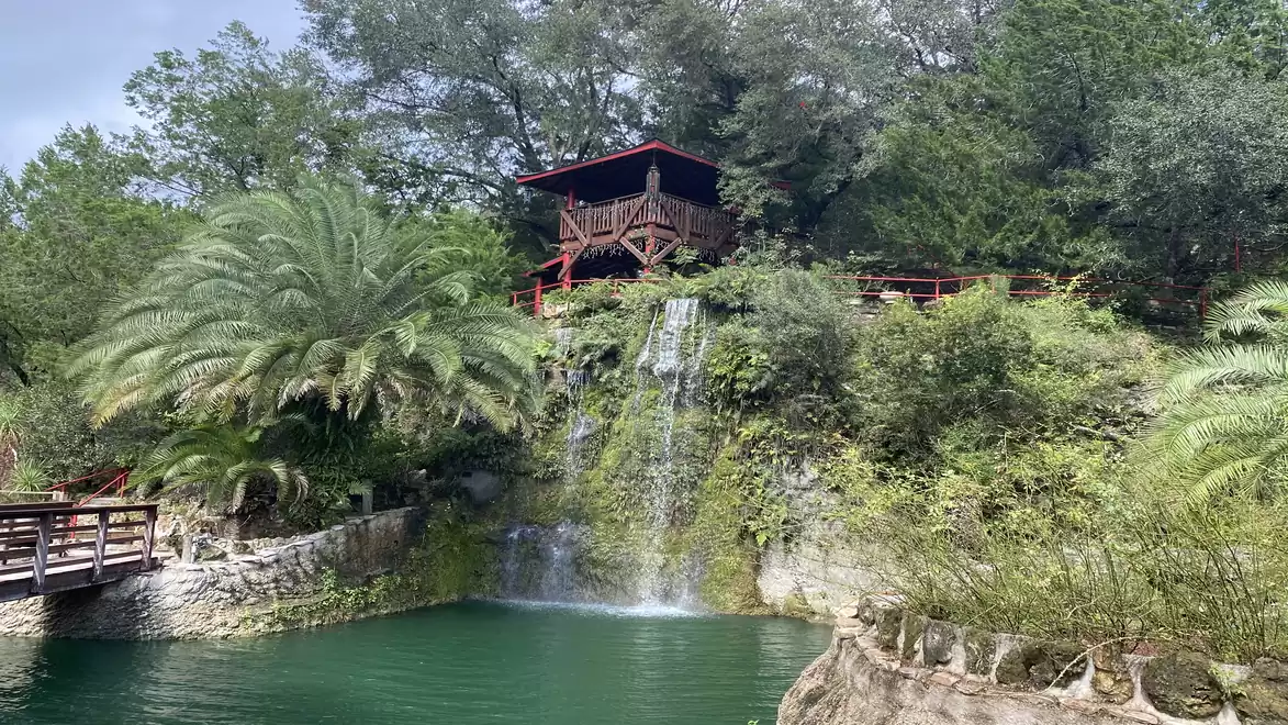 A picturesque landscape featuring a small waterfall flowing from a lush cliff into an emerald green pond. Atop the cliff is a small wooden house, surrounded by dense trees and palms.