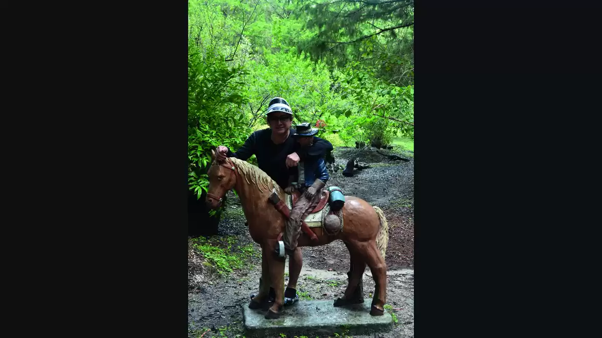 Two people on a wooden horse in the woods, surrounded by lush greenery.