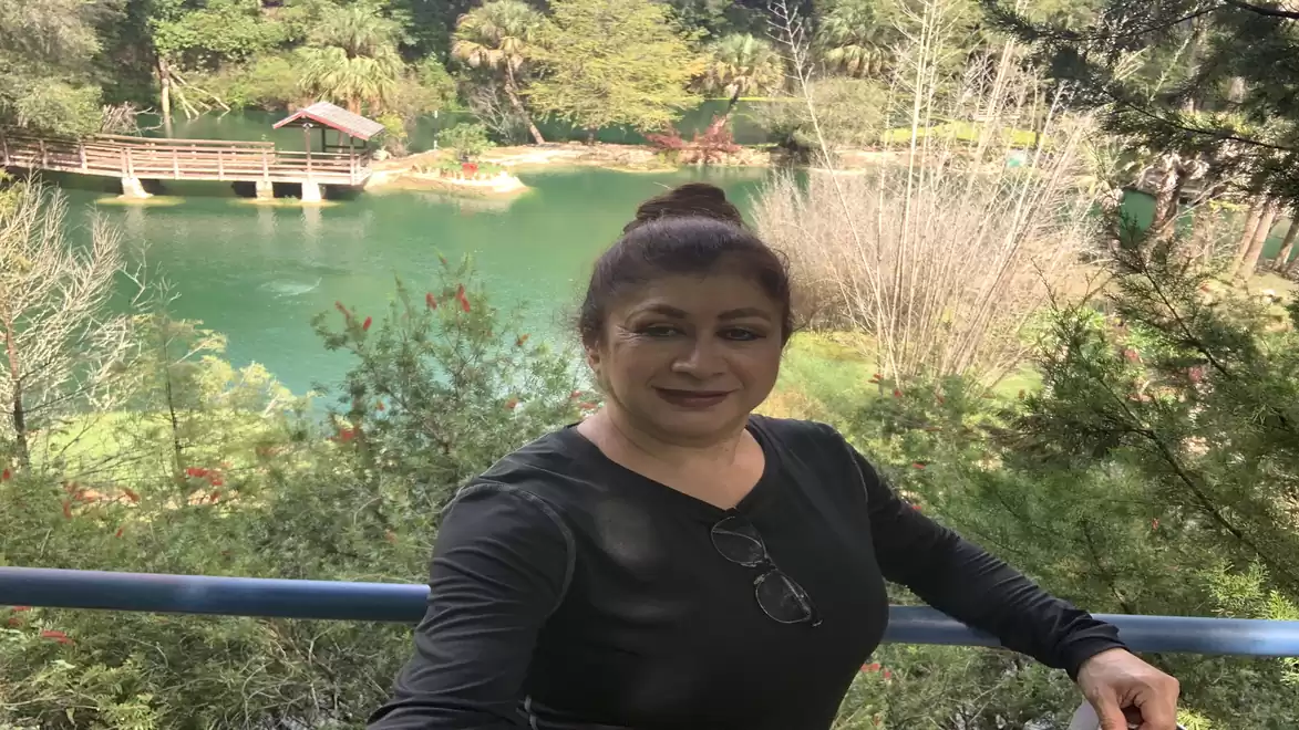 A woman in a black top takes a selfie in front of a scenic lake with green water and a wooden bridge in the background, surrounded by lush vegetation.