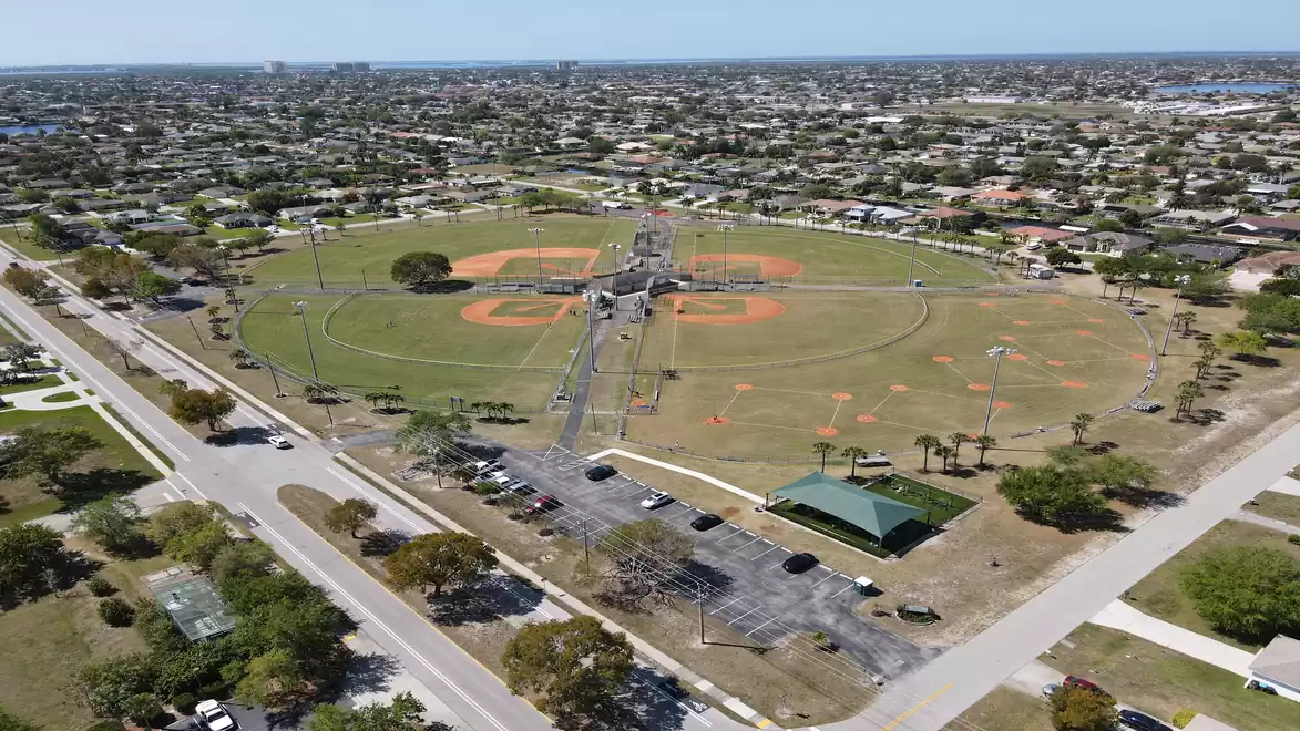 Aerial view of a baseball field located in a sprawling residential area. The field consists of several diamonds, surrounded by streets and parking lots.