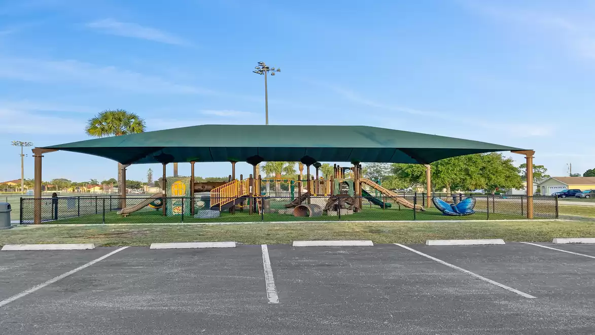 Covered playground with play equipment on a green surface, surrounded by a fence under a blue sky.