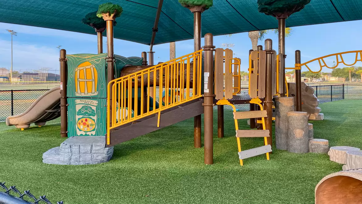 A modern playground with a slide and tree-themed elements on artificial grass under a green sunshade.