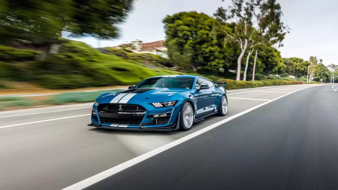 Blue Ford Shelby with white stripes driving on a road, surrounded by trees.