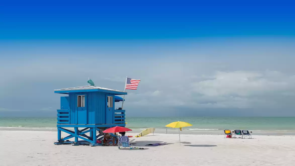 Beach with blue lifeguard tower, American flag, colorful umbrellas, and lounge chairs.