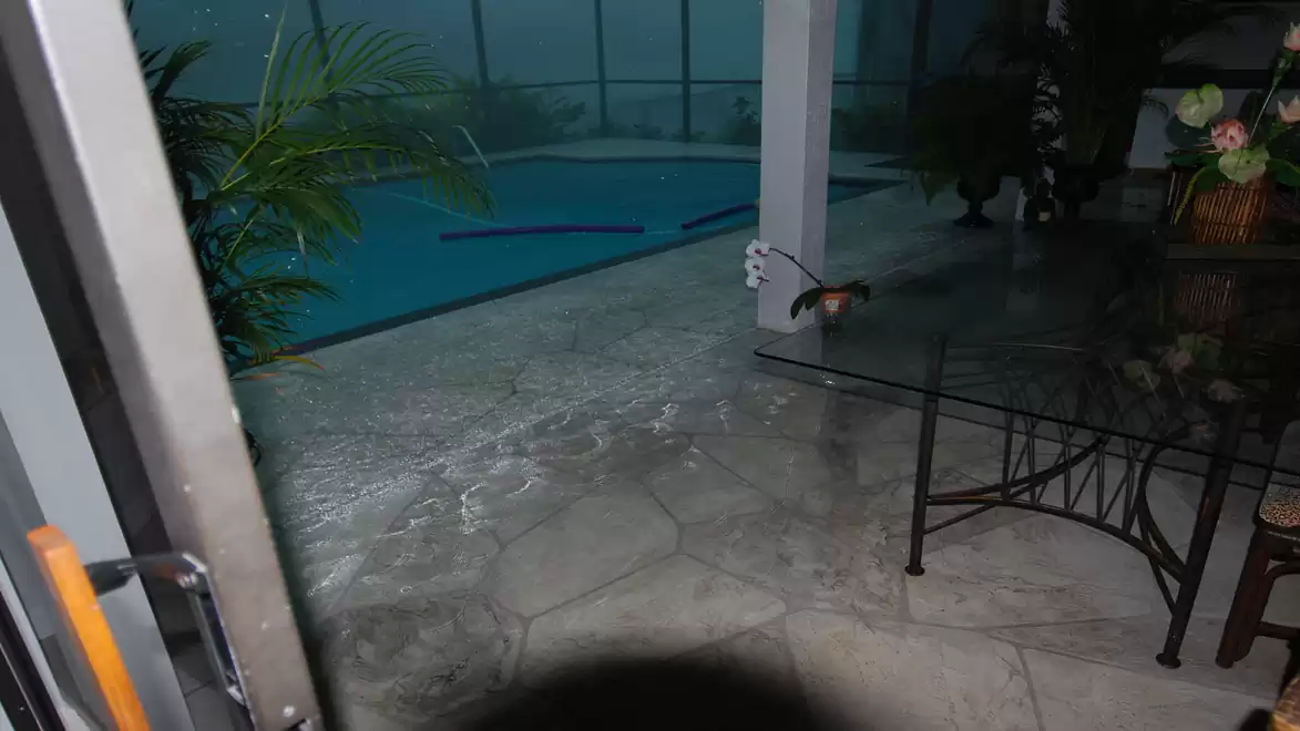 View of a covered patio overlooking an outdoor swimming pool at night. There are plants and a table in the foreground.
