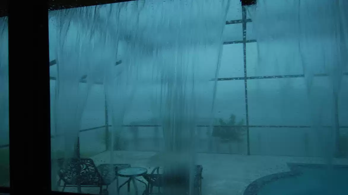 Stormy weather with heavy rain on a terrace with a pool and furniture behind a misty window.