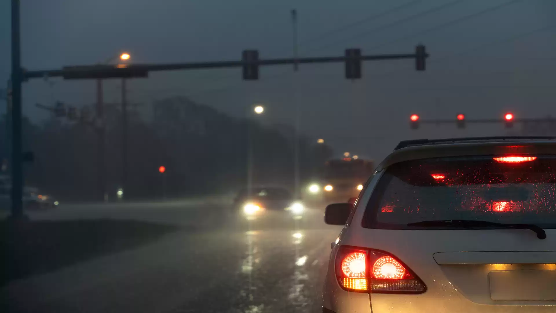 Evening traffic in rain and darkness with red taillights of a car in the foreground and blurred lights in the background at a traffic light intersection.
