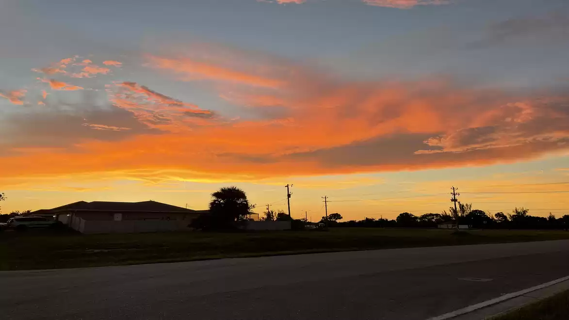An impressive sunset with bright orange clouds over a road and silhouettes of buildings and trees.