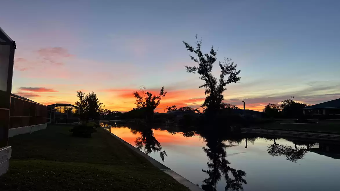 Picturesque sunset over a tranquil canal with trees and buildings reflecting in the water, featuring vibrant colors in the sky.