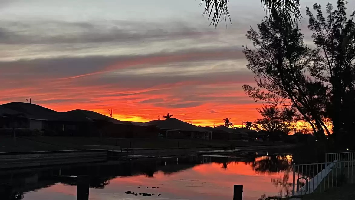 A picturesque sunset reflects in a calm river, surrounded by trees and houses.