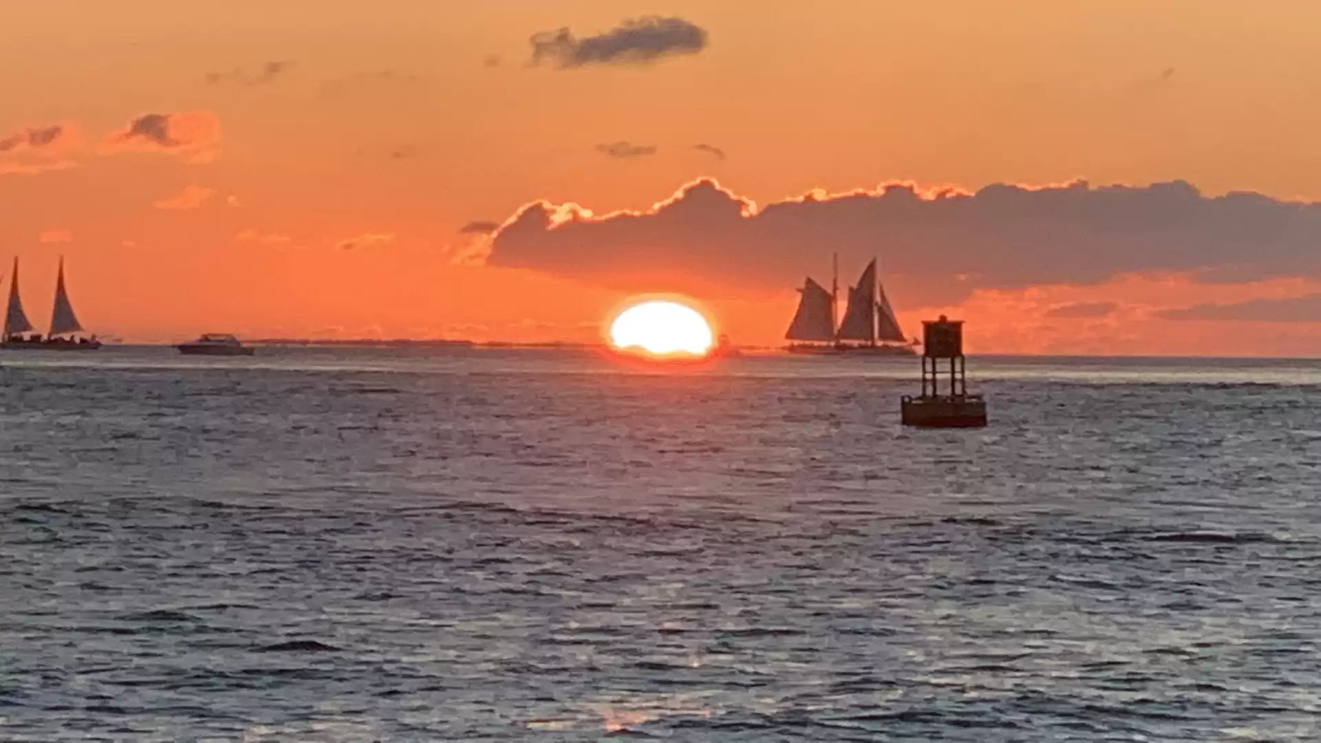 A picturesque sunset over the sea with several sailboats and a buoy in the foreground.
