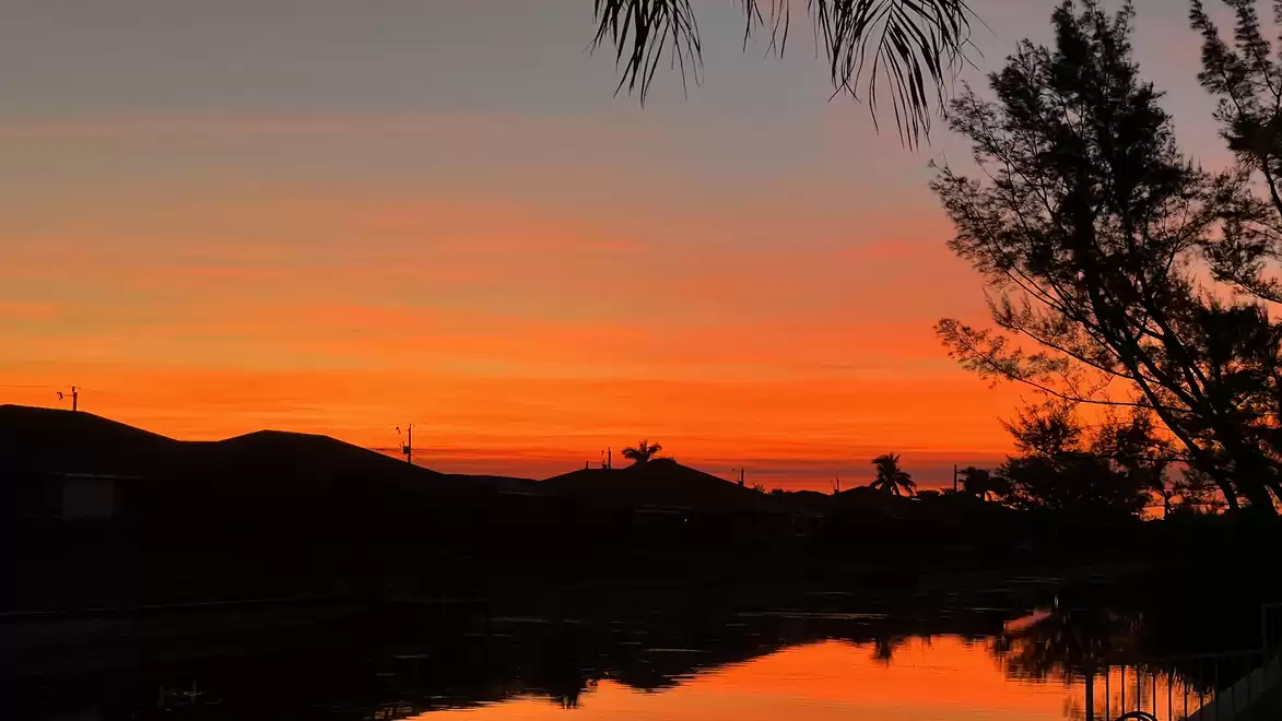 A beautiful sunset over a mountain silhouette with trees in the foreground and a calm body of water reflecting the sky's colors.