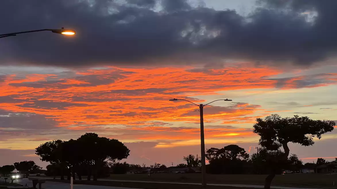 A vibrant sunset with orange clouds over a road, illuminated by streetlights and surrounded by silhouettes of trees.