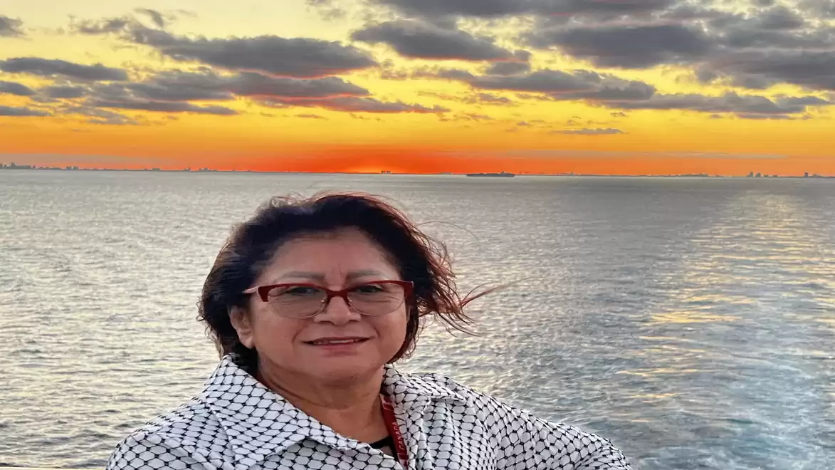 Woman with glasses and checkered shirt smiling in front of a scenic sunset over the sea.
