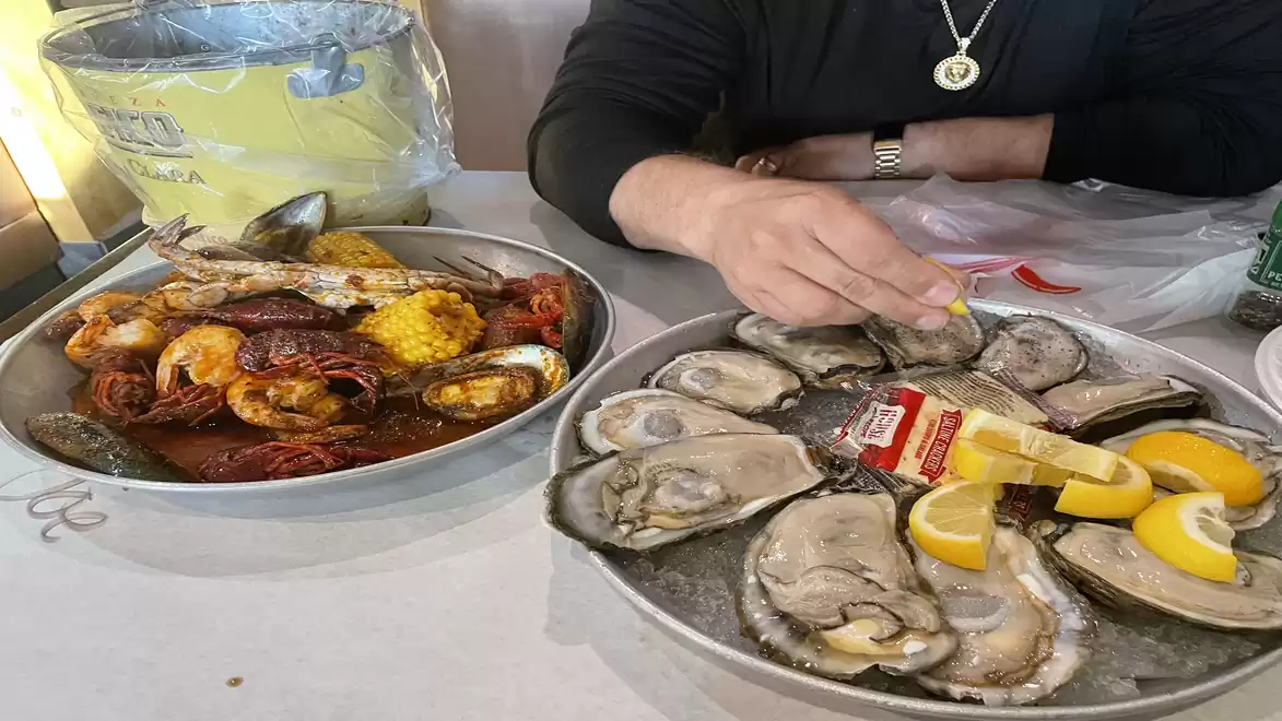 A table with two plates, one filled with seafood like mussels and crustaceans with corn and lemon slices, the other with raw oysters and lemon slices. A hand is reaching for one of the oysters.