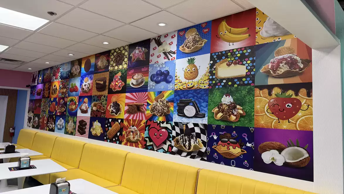 A bright wall in a café is decorated with colorful images of various ice creams and desserts. The yellow bench complements the cheerful space.
